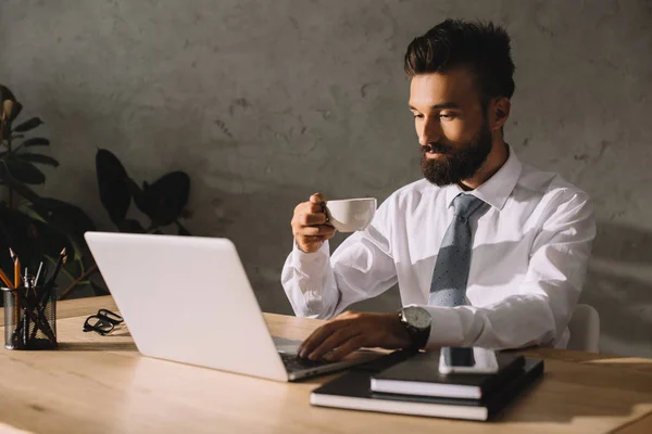 Bonito empresário segurando xícara de café usando laptop no local de trabalho — Fotografia de Stock