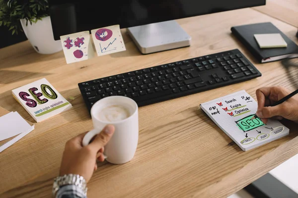 Schnappschuss eines SEO-Managers beim Schreiben von Notizen mit Tasse Kaffee und Computer am Arbeitsplatz — Stockfoto