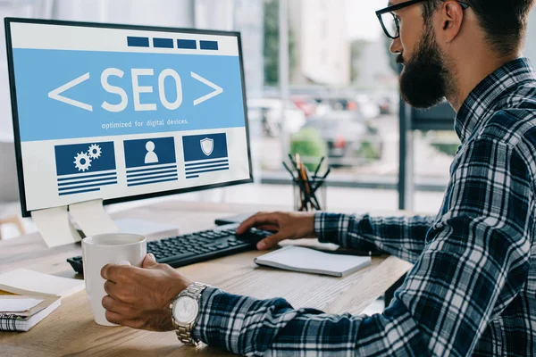 Young successful developer sitting at workplace with SEO sign on computer screen — Stock Photo