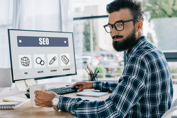 Joven gerente de SEO exitoso sentado en el lugar de trabajo con la computadora y mirando a la cámara - foto de stock