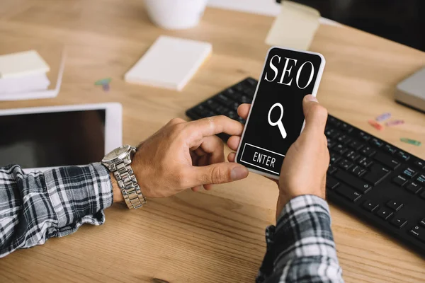 Schnappschuss von Entwickler mit Smartphone mit SEO-Zeichen am Arbeitsplatz — Stockfoto