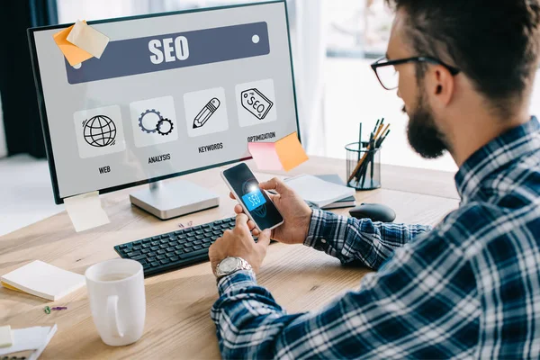 Handsome young seo manager using smartphone and computer at workplace — Stock Photo