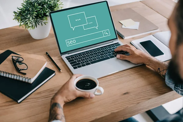 Cropped view of seo manager using laptop and holding cup of coffee at workspace — Stock Photo