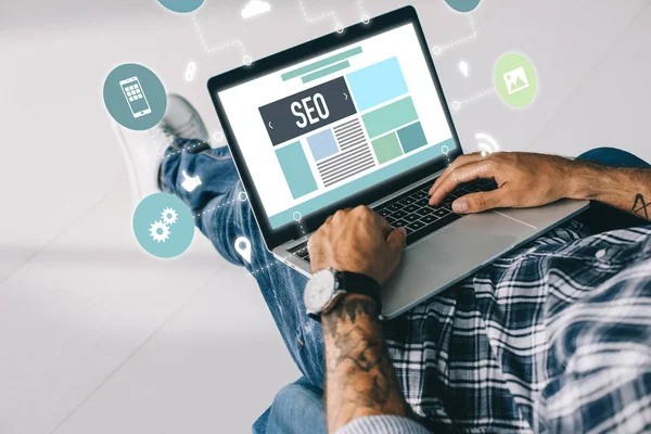 Cropped view of tattooed developer working on laptop with SEO icons — Stock Photo