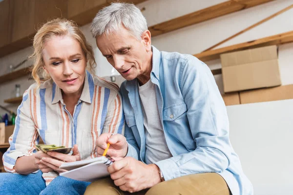 Pareja de ancianos enfocados contando dinero y tomando notas en el cuaderno durante la reubicación - foto de stock