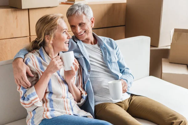 Heureux couple de personnes âgées buvant du thé et se souriant tout en étant assis sur le canapé pendant la réinstallation — Photo de stock