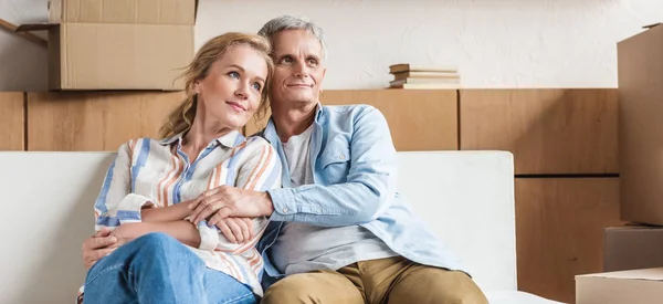 Feliz pareja de ancianos abrazando y mirando hacia otro lado mientras se sientan juntos en el sofá en la nueva casa - foto de stock