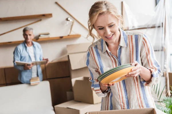 Lächelnde Frau, die Teller einpackt, während Mann mit Büchern beim Umzug zurückbleibt — Stockfoto