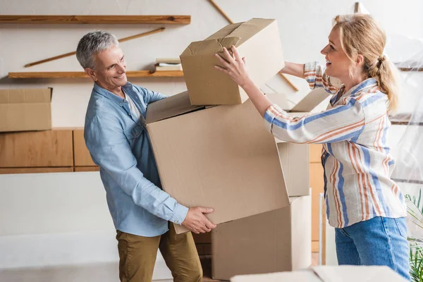 Vista lateral de feliz pareja de ancianos sosteniendo cajas de cartón mientras se muda a casa - foto de stock