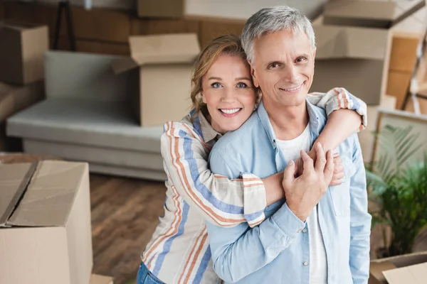 Feliz casal de idosos abraçando e sorrindo para a câmera enquanto se muda para casa — Fotografia de Stock