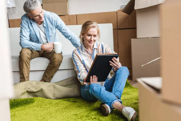 Sonriente pareja de ancianos mirando álbum de fotos mientras se sienta entre cajas de cartón en casa nueva - foto de stock