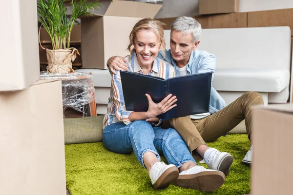 Glückliches Senioren-Paar schaut auf Fotoalbum, während es auf Teppich im neuen Haus sitzt — Stockfoto