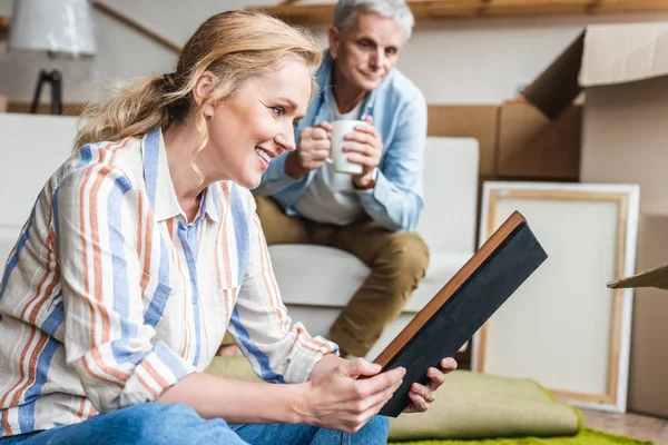 Smiling elderly woman holding photo album while relocating with husband in new apartment — Stock Photo