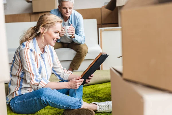 Happy elderly woman holding photo album while relocating with husband in new house — Stock Photo
