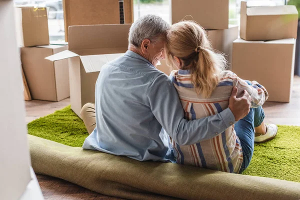 Vista trasera de la feliz pareja de ancianos abrazándose mientras está sentado en la alfombra en un nuevo hogar - foto de stock