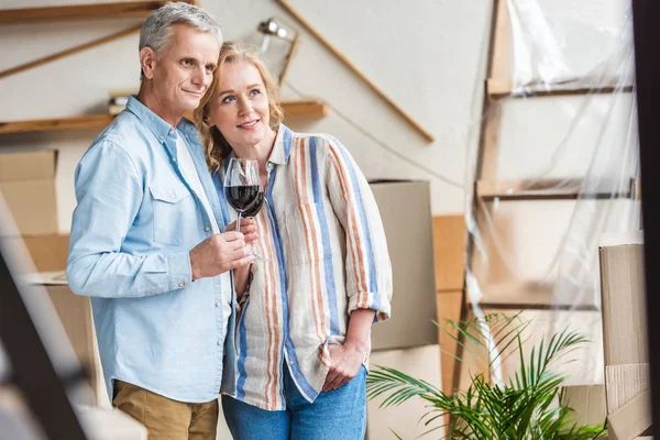 Feliz pareja de ancianos sosteniendo vasos de vino y mirando hacia otro lado mientras se muda a casa - foto de stock