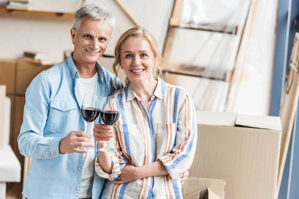 Счастливая пожилая пара держит бокалы вина и улыбается перед камерой в новой квартире — стоковое фото