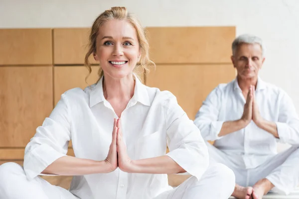 Beautiful elderly woman performing namaste gesture and smiling at camera while practicing yoga with husband — Stock Photo