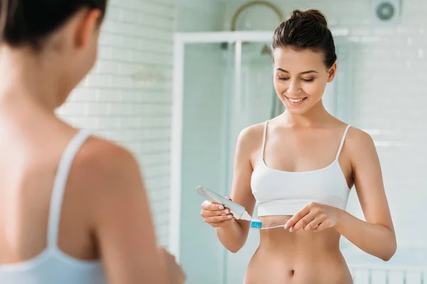 Belle fille souriante tenant brosse à dents et dentifrice au miroir dans la salle de bain — Photo de stock