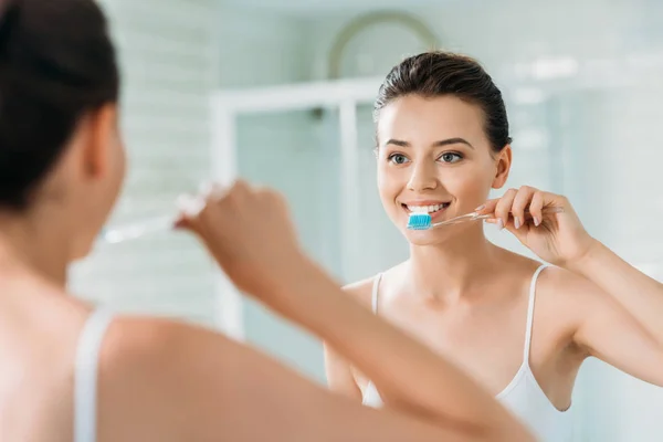 Hermosa chica sonriente cepillarse los dientes en el espejo en el baño - foto de stock