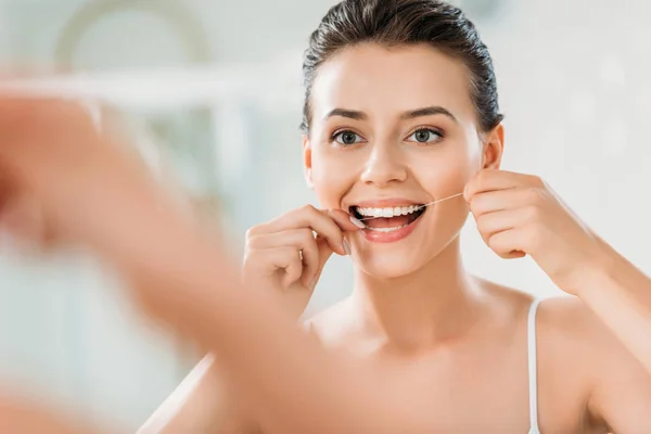 Selective focus of beautiful smiling girl using dental floss in bathroom — Stock Photo