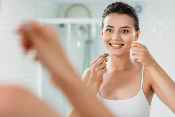 Sorridente giovane donna in possesso di filo interdentale e guardando specchio in bagno — Foto stock