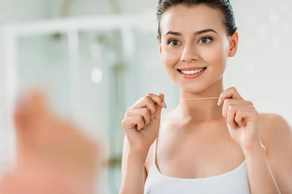 Beautiful smiling girl holding dental floss and looking at mirror in bathroom — Stock Photo