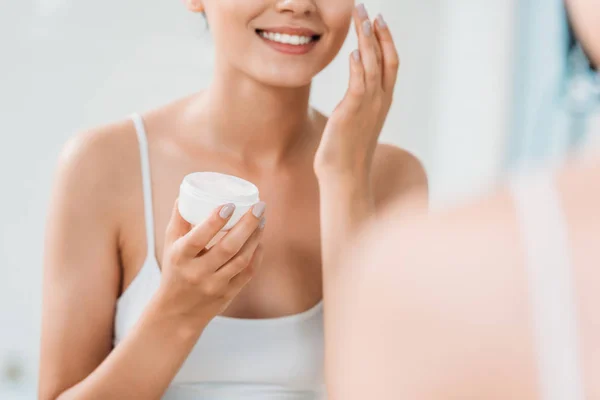 Cropped shot of smiling girl holding container and applying face cream at mirror in bathroom — Stock Photo
