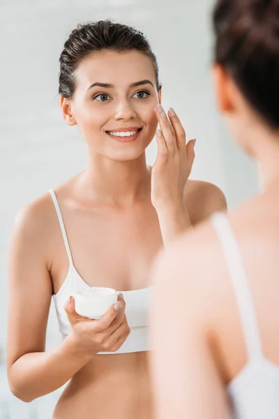 Bela sorrindo jovem mulher aplicando creme facial e olhando para o espelho no banheiro — Fotografia de Stock