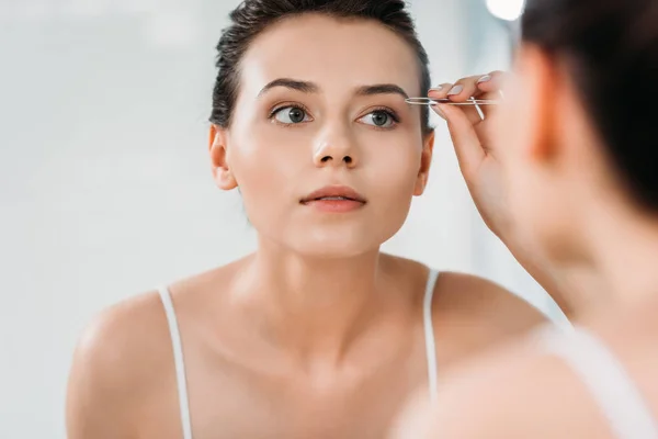 Messa a fuoco selettiva di ragazza correggere le sopracciglia con pinzette e guardando lo specchio in bagno — Foto stock