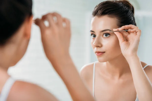 Chica atractiva corrigiendo las cejas con pinzas y mirando el espejo en el baño - foto de stock