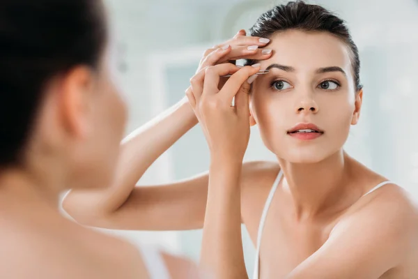 Bella ragazza correggere le sopracciglia con pinzette e guardando lo specchio in bagno — Foto stock