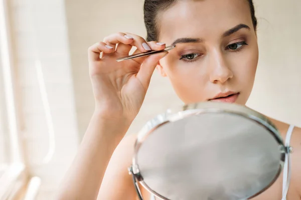 Attractive woman correcting eyebrows with tweezers and looking at mirror — Stock Photo