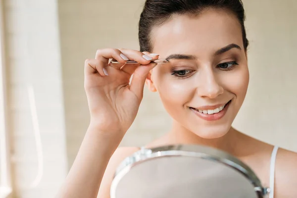 Усміхнена молода жінка виправляє брови пінцетом і дивиться на дзеркало — стокове фото
