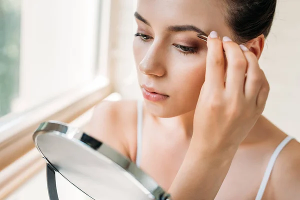 Mujer joven corrigiendo las cejas con pinzas y mirando al espejo - foto de stock