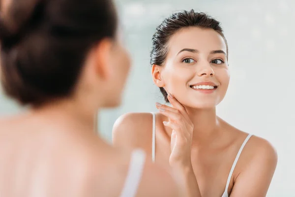 Hermosa mujer joven sonriente tocando la piel y mirando el espejo en el baño - foto de stock