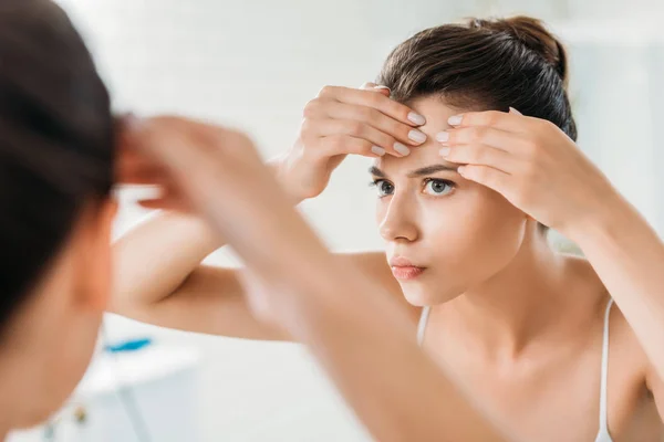 Foco seletivo da jovem mulher verificando a pele facial e olhando para o espelho no banheiro — Fotografia de Stock