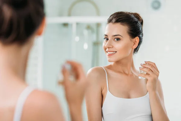Bella giovane donna in possesso di bottiglia di profumo e guardando specchio in bagno — Foto stock