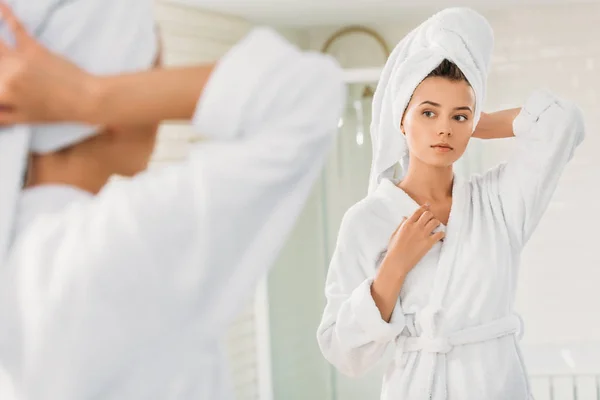 Hermosa mujer joven en albornoz y toalla en la cabeza mirando el espejo en el baño - foto de stock