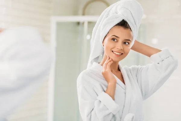 Hermosa mujer joven sonriente en albornoz y toalla en la cabeza mirando el espejo en el baño - foto de stock