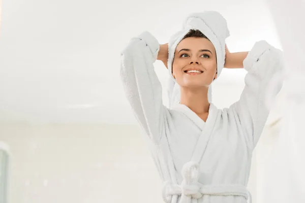Blick auf das schöne lächelnde Mädchen im Bademantel und Handtuch auf dem Kopf, das im Badezimmer wegschaut — Stockfoto
