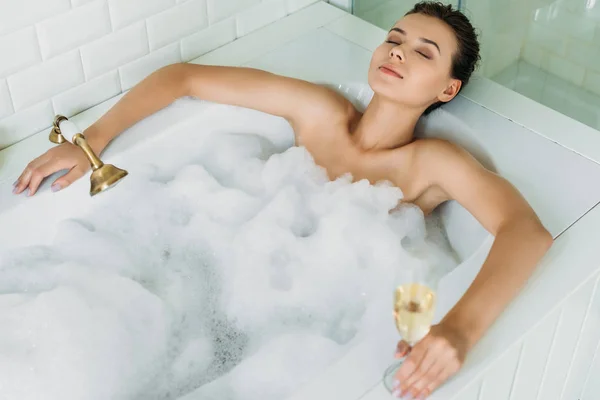 Vue grand angle de belle jeune femme aux yeux fermés tenant un verre de champagne et se relaxant dans une baignoire en mousse — Photo de stock