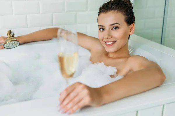 Bela jovem segurando copo de champanhe e sorrindo para a câmera no banheiro — Fotografia de Stock
