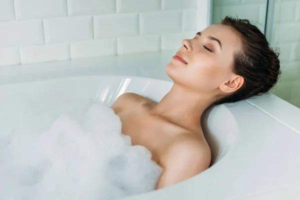 Belle jeune femme avec les yeux fermés relaxant dans la baignoire avec mousse — Photo de stock