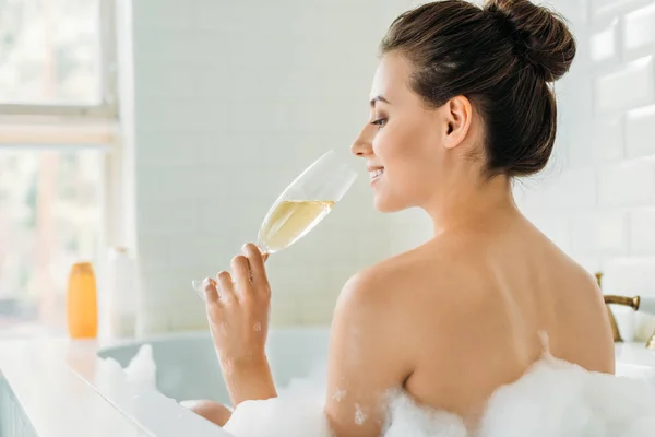 Vista trasera de hermosa chica sonriente bebiendo vino en bañera con espuma - foto de stock