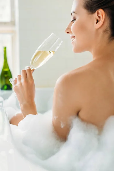 Beautiful smiling young woman drinking champagne in bathtub with foam — Stock Photo