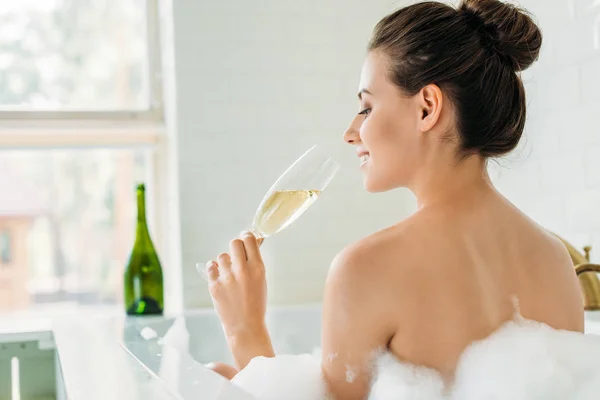 Back view of smiling young woman drinking champagne in bathtub with foam — Stock Photo