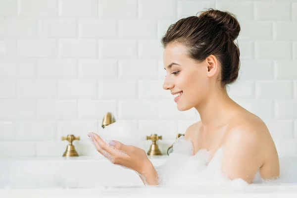 Vista lateral de hermosa chica sonriente relajándose en la bañera con espuma - foto de stock