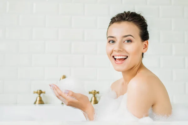 Hermosa joven sentada en la bañera con espuma y sonriendo a la cámara - foto de stock