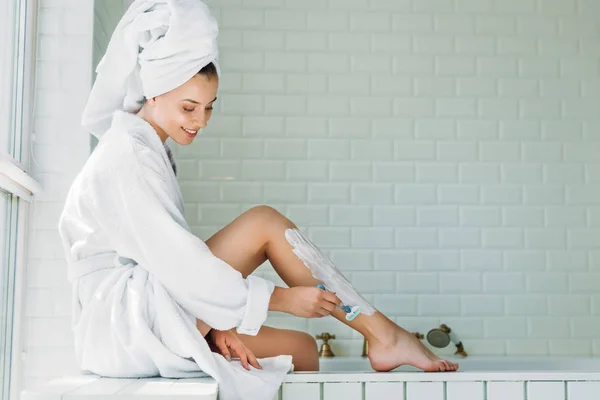 Красивая улыбающаяся молодая женщина бреет ноги бритвой в ванной комнате — стоковое фото
